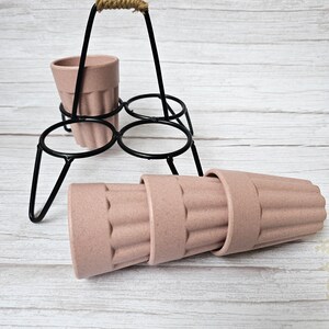 May include: A set of four pink, ribbed, reusable cups with a black metal stand with a rope handle. The cups are stacked on top of each other.