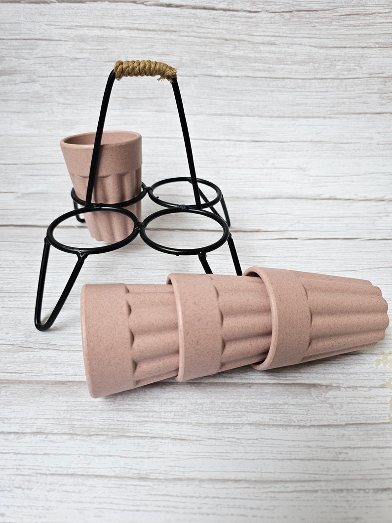 May include: A set of four pink, ribbed, reusable cups with a black metal stand with a rope handle. The cups are stacked on top of each other.