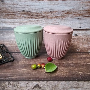 May include: Two lidded cups, one light green and one pink, sit on a wooden surface. The cups have a ribbed design and a matching lid. Decorative fruit and a green leaf are placed near the cups.