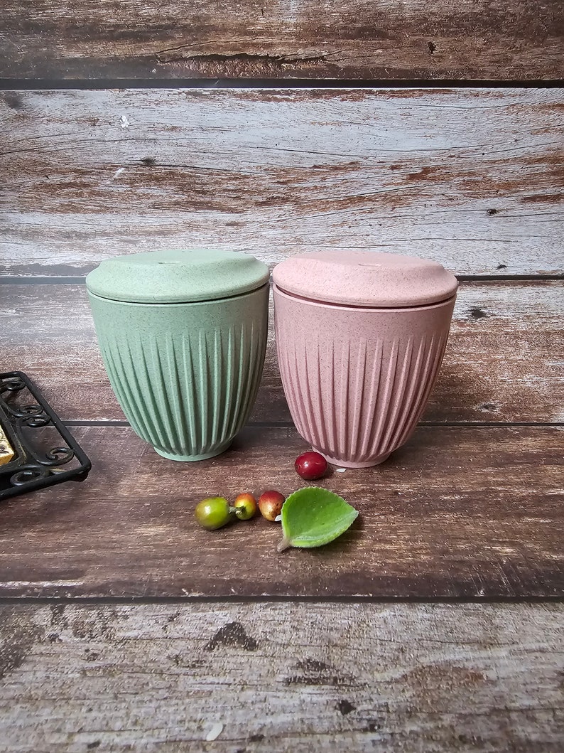 May include: Two lidded cups, one light green and one pink, sit on a wooden surface. The cups have a ribbed design and a matching lid. Decorative fruit and a green leaf are placed near the cups.