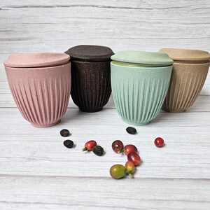 May include: Four reusable coffee cups in pink, brown, mint green, and tan, each with a matching lid. The cups have a ribbed design and are displayed with coffee beans and berries on a light wood-grain surface.