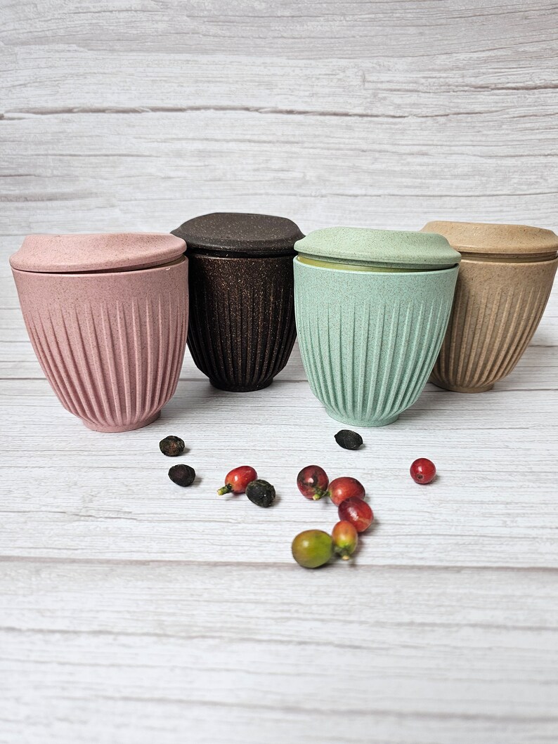 May include: Four reusable coffee cups in pink, brown, mint green, and tan, each with a matching lid. The cups have a ribbed design and are displayed with coffee beans and berries on a light wood-grain surface.