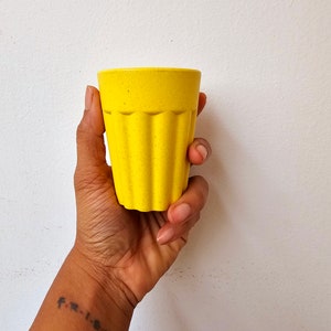 May include: A yellow, textured, ceramic drinking glass with a ribbed design. The glass is being held in a person's hand.