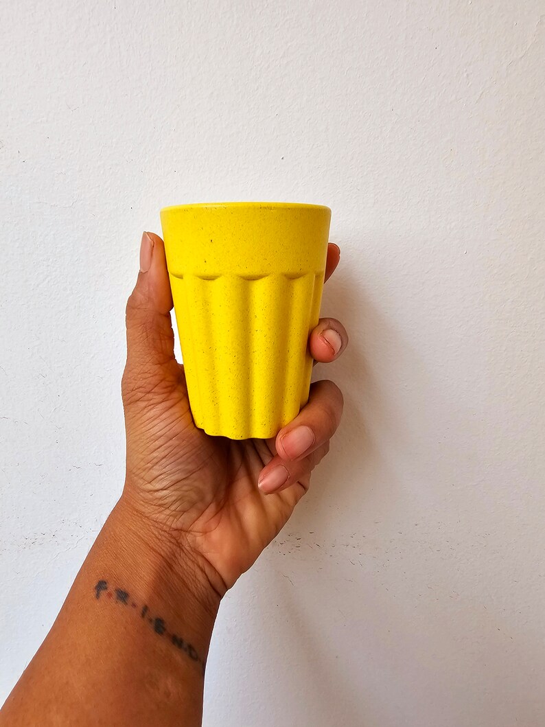 May include: A yellow, textured, ceramic drinking glass with a ribbed design. The glass is being held in a person's hand.