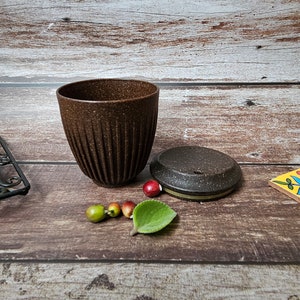 May include: A brown ceramic cup with a textured surface and vertical grooves. The cup is next to a matching lid and several small decorative fruits and a leaf. The items are arranged on a wooden surface.