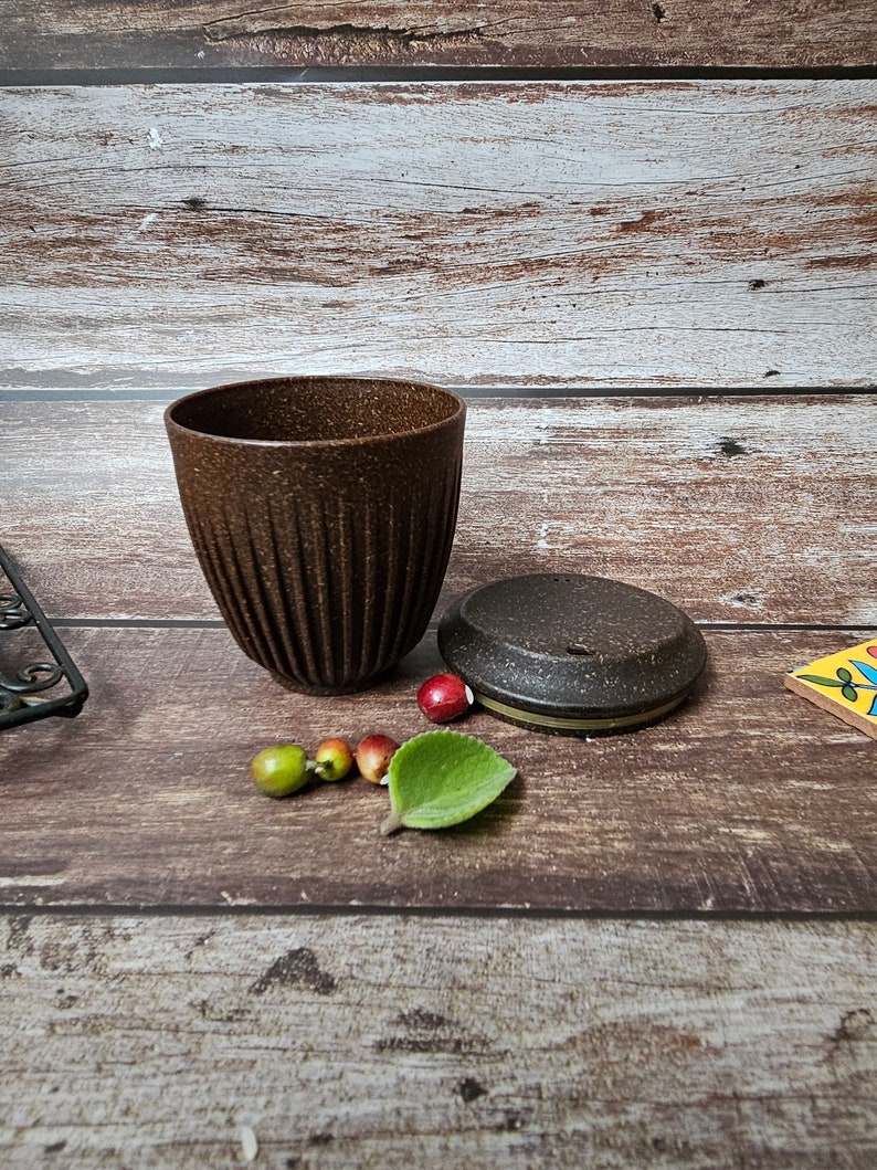 May include: A brown ceramic cup with a textured surface and vertical grooves. The cup is next to a matching lid and several small decorative fruits and a leaf. The items are arranged on a wooden surface.