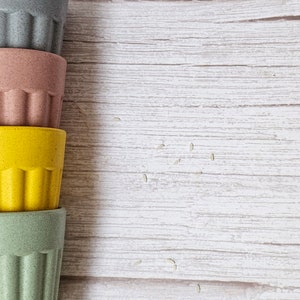 May include: Four stacked reusable cups made of bamboo. The cups are in shades of green, pink, yellow, and gray. The cups are on a white wooden surface with scattered white rice grains.