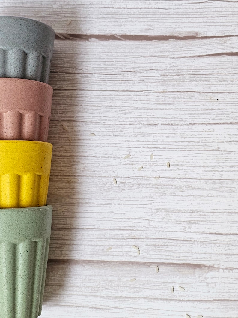 May include: Four stacked reusable cups made of bamboo. The cups are in shades of green, pink, yellow, and gray. The cups are on a white wooden surface with scattered white rice grains.