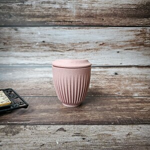 May include: A pink reusable coffee cup with a ridged design. The cup has a matching lid and is made of a durable material. The cup is designed for hot or cold beverages and is a sustainable alternative to disposable cups.