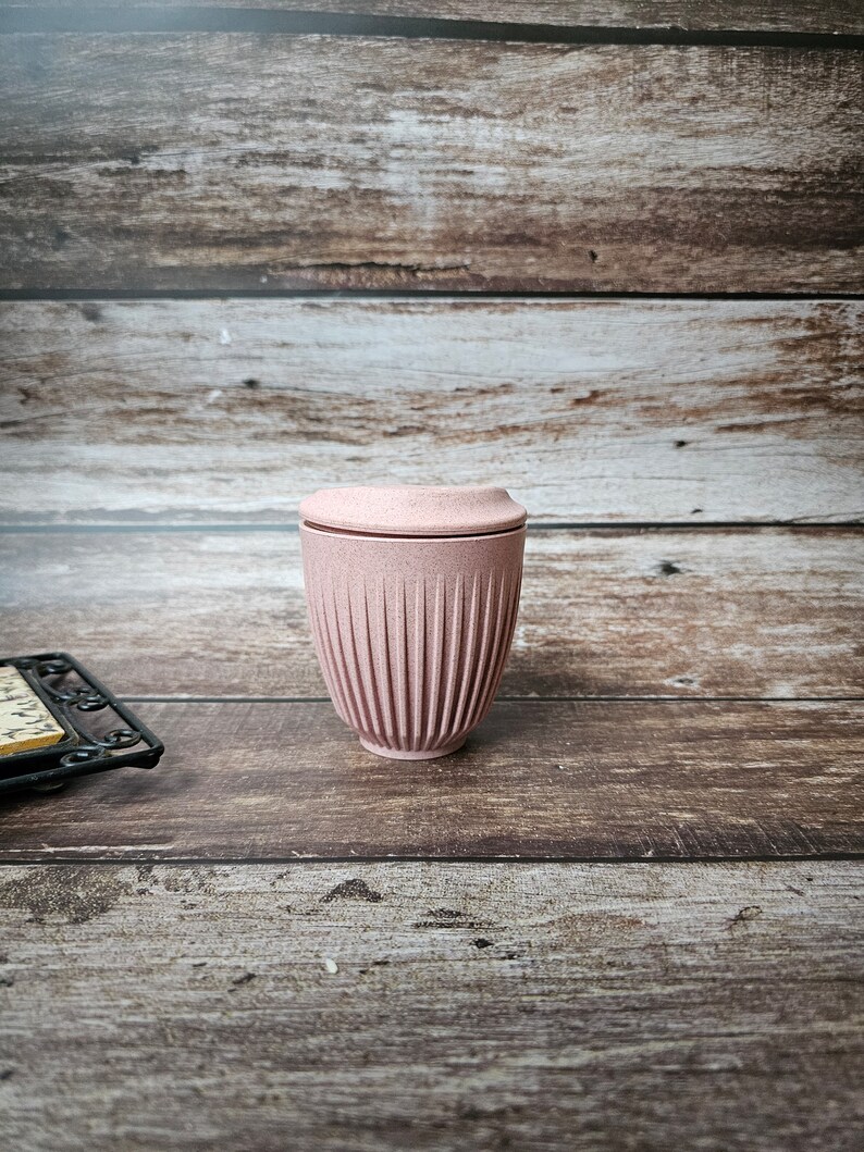 May include: A pink reusable coffee cup with a ridged design. The cup has a matching lid and is made of a durable material. The cup is designed for hot or cold beverages and is a sustainable alternative to disposable cups.