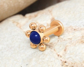 Pode incluir: Um piercing labret em tom dourado com uma pedra central circular azul-escura rodeada por seis pequenas esferas douradas polidas dispostas em forma de flor. A peça possui um disco traseiro plano e uma haste reta e lisa.