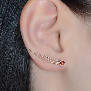 Puede incluir: Un par de pendientes de aro de oro con una pequeña piedra roja. Los pendientes están diseñados para curvarse a lo largo de la oreja.