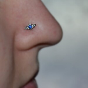 Op de afbeelding: Een close-up van een neuspiercing met een klein blauw steentje en zilveren accenten.