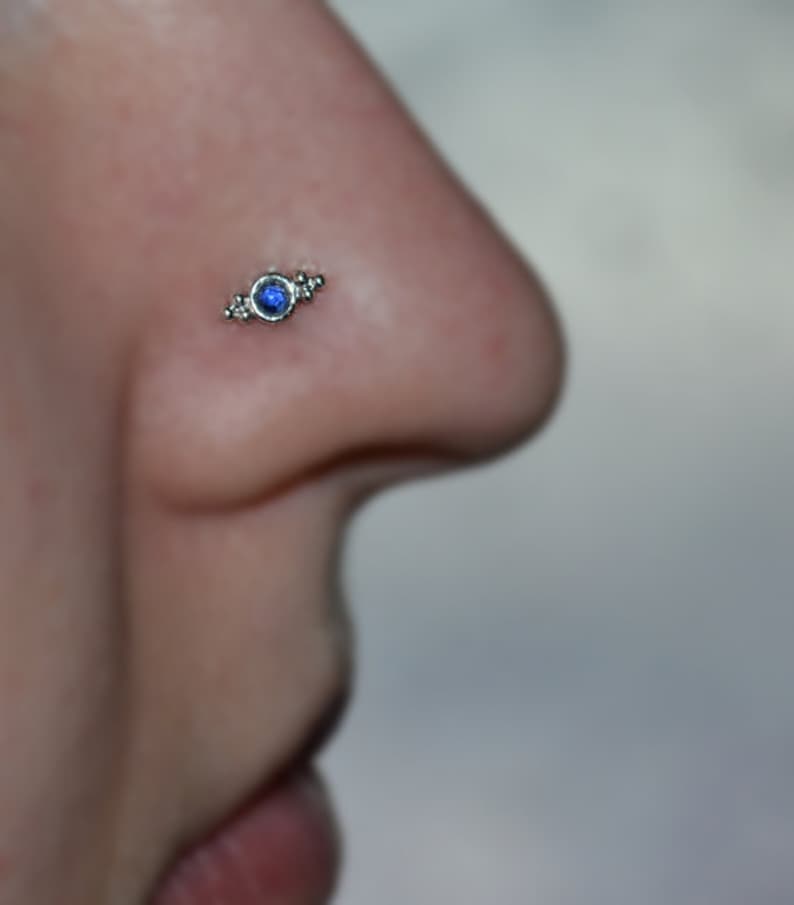 Op de afbeelding: Een close-up van een neuspiercing met een klein blauw steentje en zilveren accenten.
