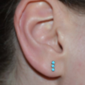 May include: A close-up of a person's ear with a turquoise and gold stud earring. The earring has three turquoise stones in a vertical line.