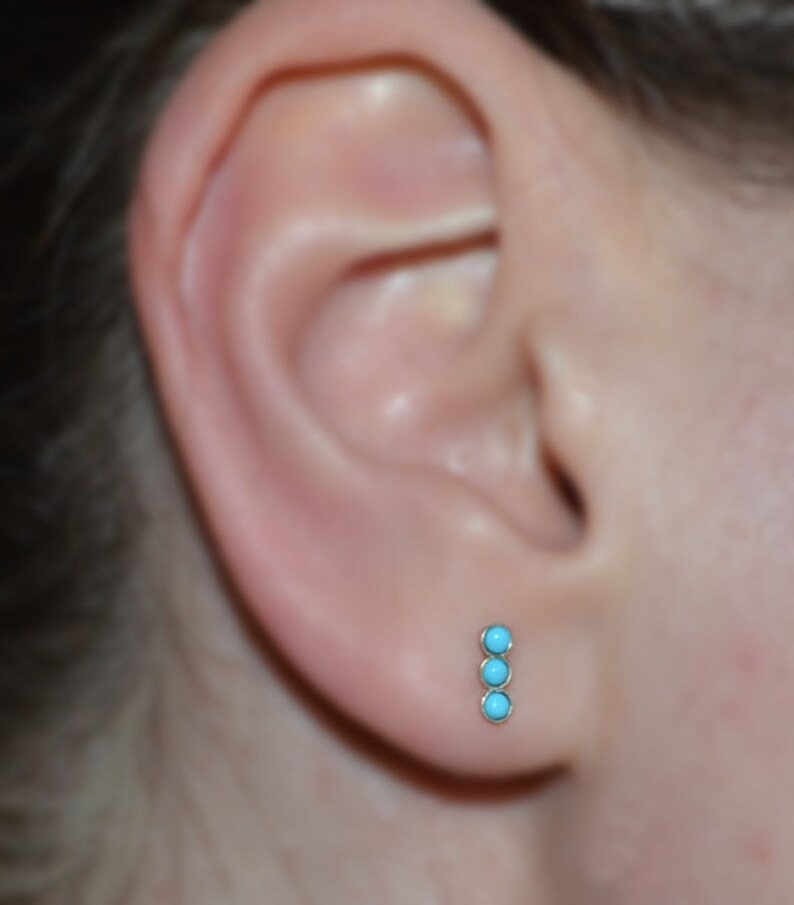 May include: A close-up of a person's ear with a turquoise and gold stud earring. The earring has three turquoise stones in a vertical line.