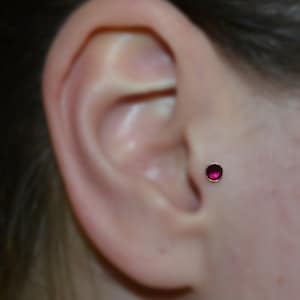 Peut inclure: Gros plan sur une oreille avec un petit piercing en forme de clou avec une pierre précieuse rose. Le piercing est serti dans un cadre en or.