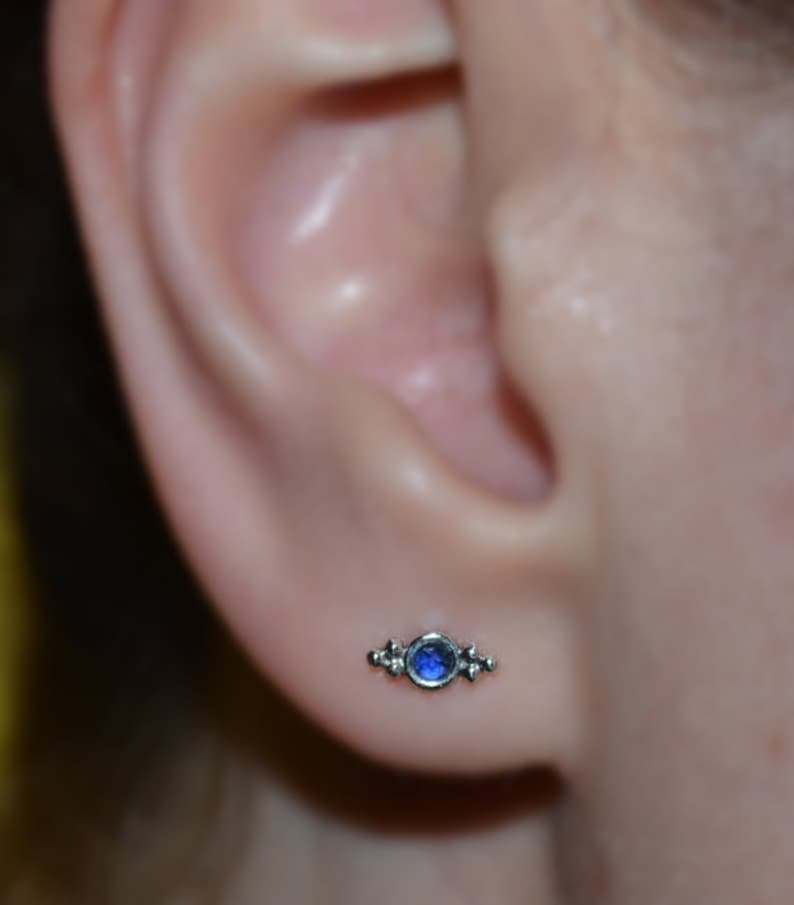 Op de afbeelding: Een zilveren oorbel met een klein blauw edelsteentje en drie kleine zilveren kralen aan weerszijden van de steen.