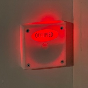 May include: A white square-shaped sign with a red light behind it. The sign says "OCCUPIED" in black letters.