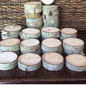 May include: A collection of 20 natural wood slices, each approximately 4 inches in diameter, with bark still attached. The wood slices are arranged in a circular pattern on a dark wooden surface.