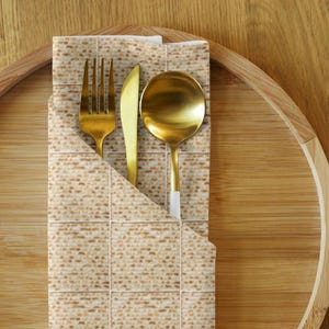 Puede incluir: Un juego de tenedor, cuchillo y cuchara dorados descansa en una servilleta con un patrón de matzá, presentado en una bandeja de madera. Los utensilios tienen un acabado brillante, y la servilleta está doblada para crear un bolsillo.