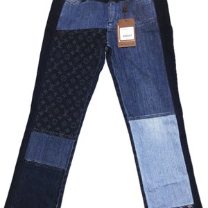 Puede incluir: Vaqueros patchwork de mezclilla con varios tonos de azul. Los vaqueros presentan una mezcla de paneles lisos y paneles con un patrón de logotipo repetido. Una etiqueta marrón cuelga de la cintura. El texto "LOUIS VUITTON" es visible en la cintura.