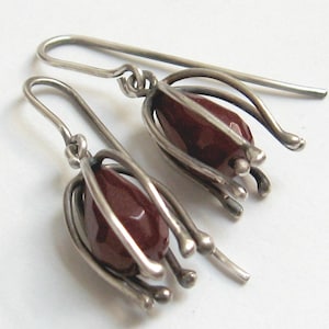 May include: A pair of silver drop earrings featuring a faceted, teardrop-shaped dark red stone encased within a cage of curved silver wires. The earrings hang from simple silver hooks and have a polished, metallic finish with a dark patina.