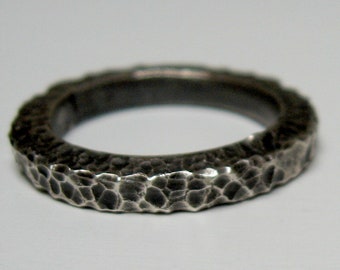 May include: A dark silver-toned ring featuring a heavily textured, hammered surface. The band has a rugged, uneven finish with deep indentations across the exterior, creating a high-contrast appearance between the dark recesses and the polished edges.