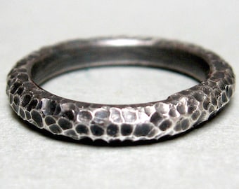 May include: A dark grey metal ring with a textured, hammered surface. The band features a series of small, irregular indentations across its entire outer circumference, creating a rugged, uneven appearance with high-contrast highlights and shadows.