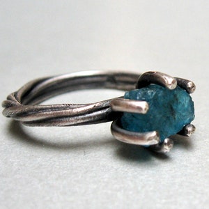 May include: A ring featuring a raw, blue, unpolished gemstone held in a four-prong setting. The band is crafted from dark, oxidized metal with a twisted, rope-like texture that wraps around the finger, providing a rugged and textured appearance.
