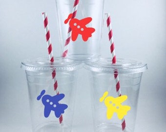 May include: Three clear plastic cups with red and white striped straws. Each cup has a paper airplane sticker with different colors: red, blue, and yellow.