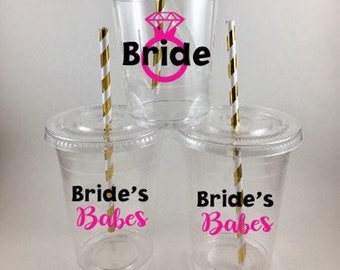 May include: Three clear plastic cups with lids and straws. The cups are decorated with black text that says "Bride" and "Bride's Babes". One cup has a pink diamond ring graphic.
