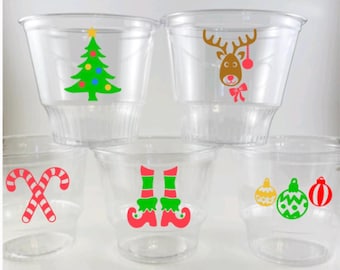 May include: Five clear plastic cups with festive Christmas designs. The designs include a Christmas tree, a reindeer, elf shoes, and two ornaments.