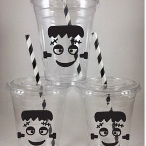 May include: Three clear plastic cups with lids, each decorated with a black Frankenstein face. The cups have black and white striped straws inserted. The cups are stacked in a pyramid shape.