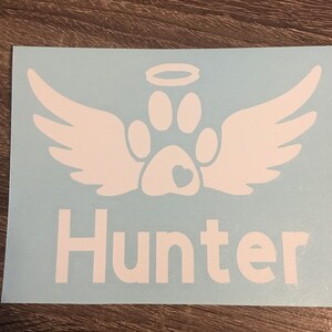 May include: White vinyl decal with a paw print with a heart inside, angel wings, and a halo above. The name "Hunter" is written below the paw print.