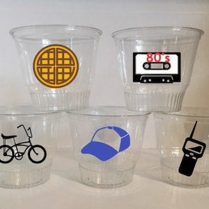 May include: Set of six clear plastic cups with various Stranger Things themed designs. The designs include a waffle, a cassette tape with the text "80's", a bicycle, a baseball cap, and a walkie talkie.