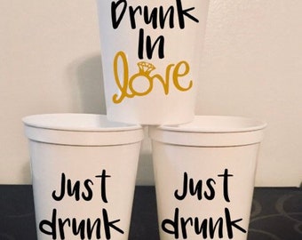 May include: Three white plastic cups with black text. The top cup says "Drunk in Love" with a gold ring design. The two bottom cups say "Just Drunk".