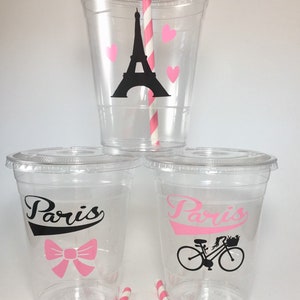 May include: Three clear plastic cups with pink and black designs. The top cup has a black silhouette of the Eiffel Tower and pink hearts. The bottom two cups have the word "Paris" in pink and black with a pink bow and a black bicycle with a basket.
