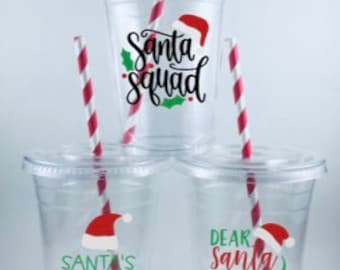 May include: Three clear plastic cups with red and white striped straws. Each cup has a different Christmas-themed design with a red Santa hat graphic. The designs read "Santa Squad", "Santa's Cookie Tester", and "Dear Santa Define Good".