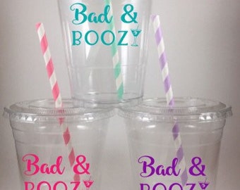May include: Three clear plastic cups with straws and the text "Bad & Boozy" printed on them. The text is in different colors on each cup: turquoise, pink, and purple.