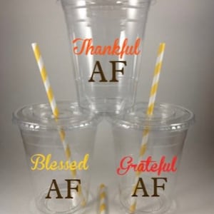 May include: Clear plastic cups with lids and yellow and white striped straws. The cups have the words "Thankful AF", "Blessed AF", and "Grateful AF" in various colors. The cups are stacked on top of each other.