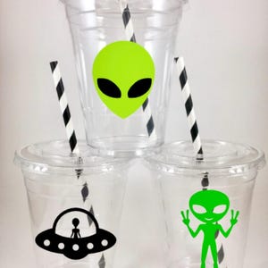 May include: Three clear plastic cups with lids and black and white striped straws. Each cup has a different alien design: a green alien head, a green alien with peace signs, and a black alien in a spaceship.