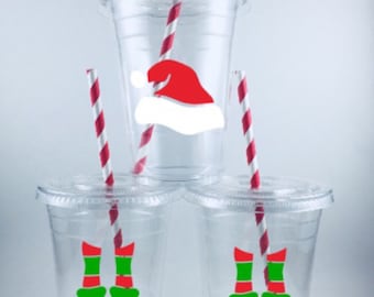 May include: Three clear plastic cups with red and white striped straws. The cups have festive Christmas designs, one with a Santa hat and two with elf shoes.