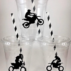 K&ouml;nnte beinhalten: Drei durchsichtige Plastikbecher mit Deckeln und schwarz-wei&szlig; gestreiften Strohhalmen. Jeder Becher hat eine schwarze Silhouette einer Person, die ein Dirtbike f&auml;hrt.