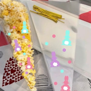 May include: Clear plastic cone-shaped bags with bunny rabbit designs in pink, purple, and blue. The bags are filled with popcorn and have gold twist ties.