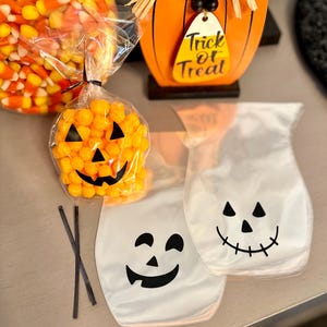 May include: Two clear plastic treat bags with black pumpkin and ghost faces printed on them. One bag is filled with orange cheese puffs. The bags are on a wooden surface with a black and orange candy corn bowl and a wooden pumpkin sign that says "Trick or Treat".