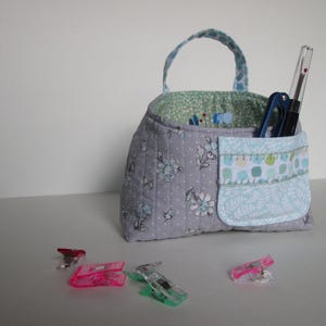 May include: A quilted fabric organizer with a handle, featuring a floral pattern in gray and blue. The organizer has a pocket and contains sewing tools. Several colorful clips are scattered in front of it.