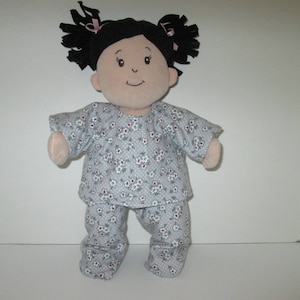 Puede incluir: Una muñeca con un pijama de estampado floral blanco y azul claro. La muñeca tiene el pelo negro en coletas con lazos rosas.