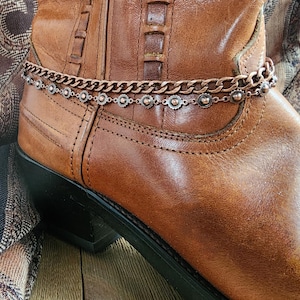 May include: A close-up of a brown leather cowboy boot with a copper-colored boot chain. The boot chain has a delicate design with small circular accents. The boot is worn and has visible stitching and a black heel.