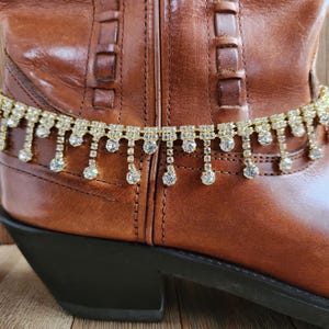May include: A gold-toned boot chain with dangling clear rhinestones is draped over a brown leather cowboy boot. The boot has a black heel and is set against a wooden background.